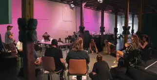 Eine Gruppe von Menschen sitzt in einem grosen Raum im Kreis zusammen. Einige Personen sitzen auf Stühlen oder im Rollstuhl, andere auf dem Boden oder auf einem Sitzsack.